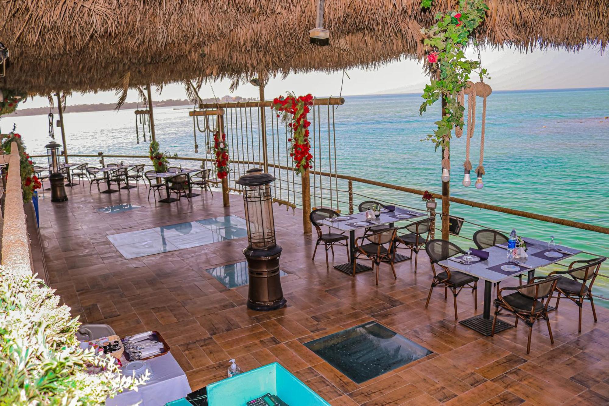 Lagoona Beach Luxury Resort And Spa Манама Экстерьер фото The photo depicts a coastal restaurant or dining area with an open-air design. It features a wooden floor and tables set for dining, surrounded by chairs. The space is partially covered by a thatched roof, providing shade while allowing for a view of