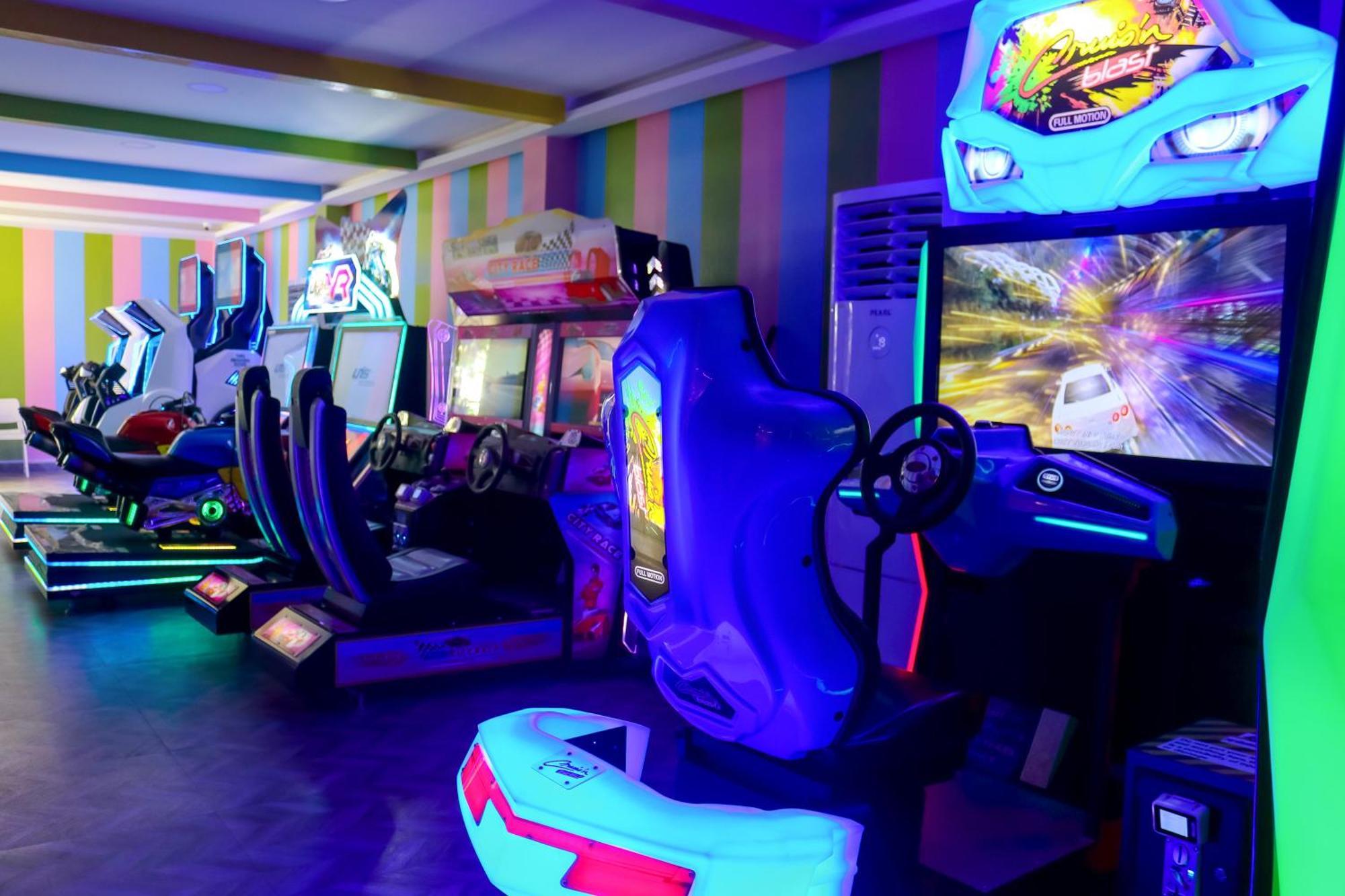 Lagoona Beach Luxury Resort And Spa Манама Экстерьер фото The photo shows an arcade area filled with various arcade machines. The setting is illuminated with colorful neon lights, creating a vibrant atmosphere. There are several racing game machines with steering wheels and seats, each accompanied by screen