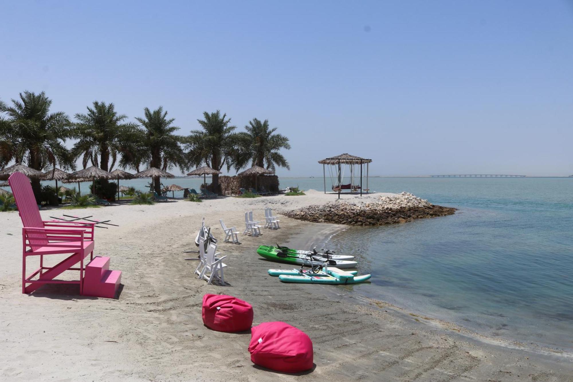 Lagoona Beach Luxury Resort And Spa Манама Экстерьер фото The photo depicts a serene beach scene with clear blue water and a sunny sky. In the foreground, there are colorful pink bean bags and a large pink chair on the sand. A few white chairs and loungers are positioned near the water's edge. To the side, 