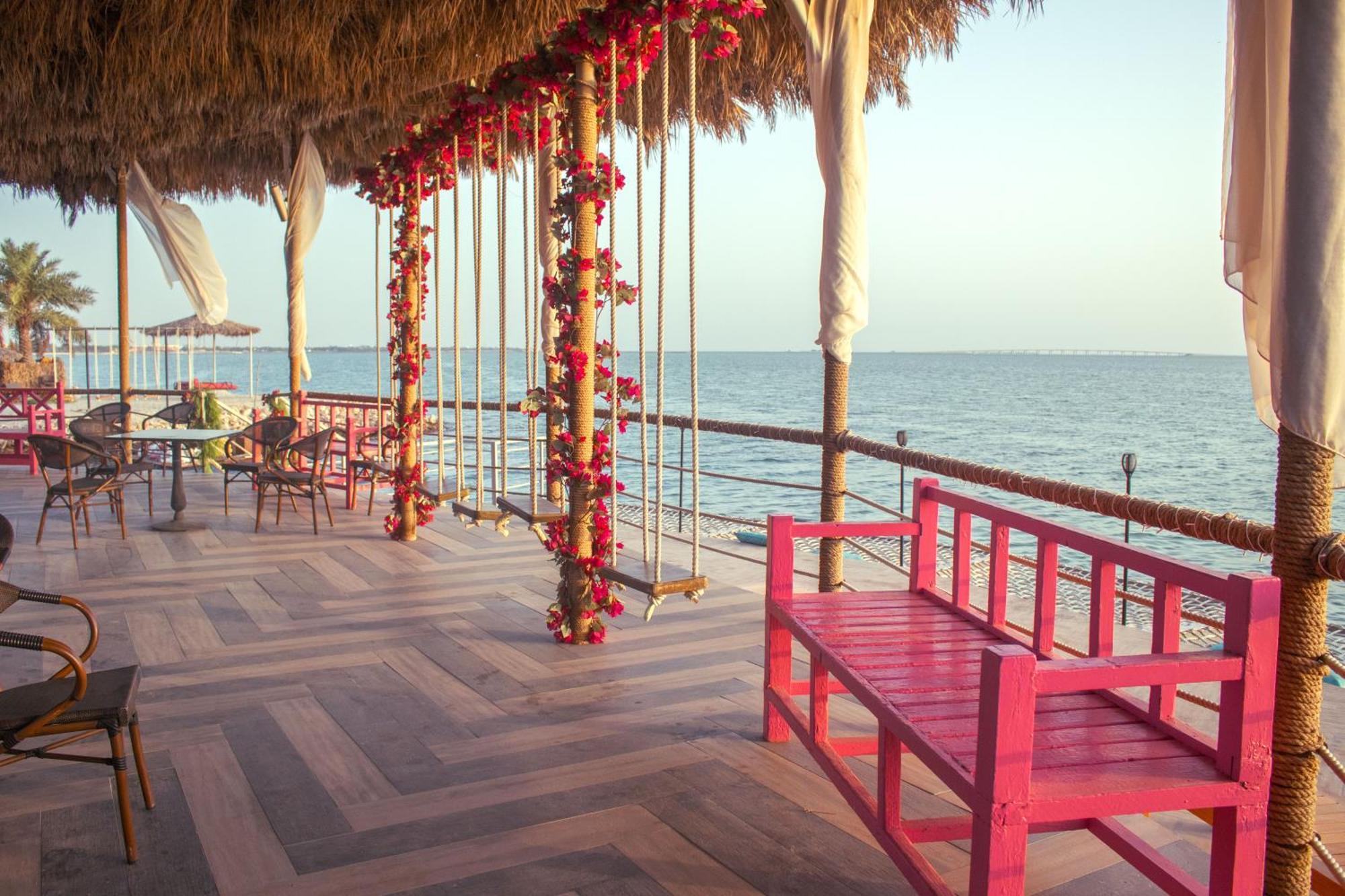 Lagoona Beach Luxury Resort And Spa Манама Экстерьер фото The photo depicts a scenic outdoor area by the water, with a wooden deck and a thatched roof. Two swings adorned with flowers are hanging from the ceiling, providing a charming touch to the space. In the foreground, there is a bright pink bench, addi