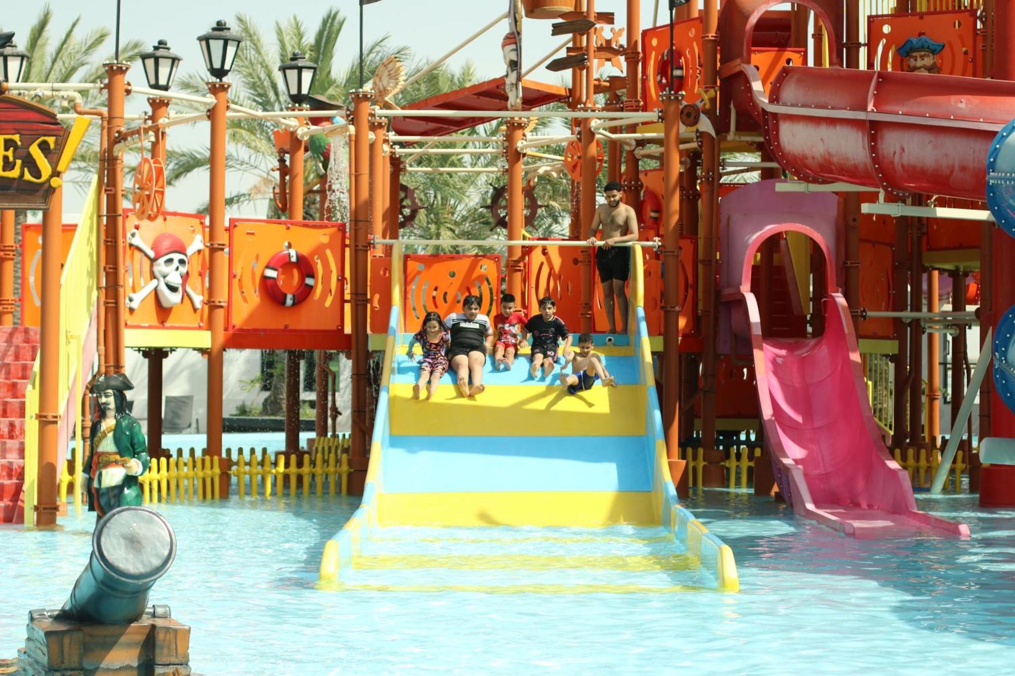 Lagoona Beach Luxury Resort And Spa Манама Экстерьер фото The photo shows a colorful water park scene featuring a play area with multiple slides. There are children enjoying themselves, sliding down bright yellow and pink slides into a pool of water. The structure has pirate-themed decorations, including a 