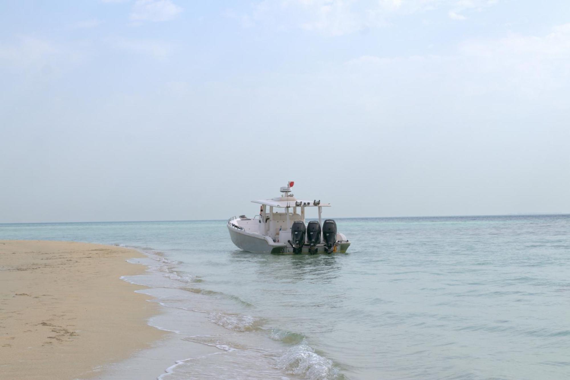 Lagoona Beach Luxury Resort And Spa Манама Экстерьер фото The photo depicts a small boat anchored near a sandy beach, partially submerged in calm, clear water. The boat is white and features three outboard motors on its stern. In the background, the sea stretches out toward the horizon under a light, cloudy