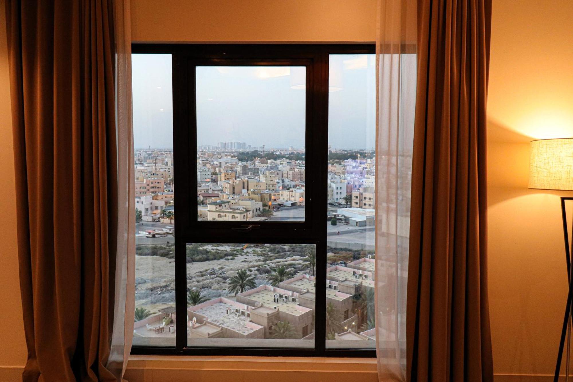 Lagoona Beach Luxury Resort And Spa Манама Экстерьер фото The photo shows a window with a black frame and sheer curtains. Through the window, there is a view of a cityscape, displaying a densely populated area with various buildings and rooftops. The sky appears slightly overcast, suggesting a muted lightin