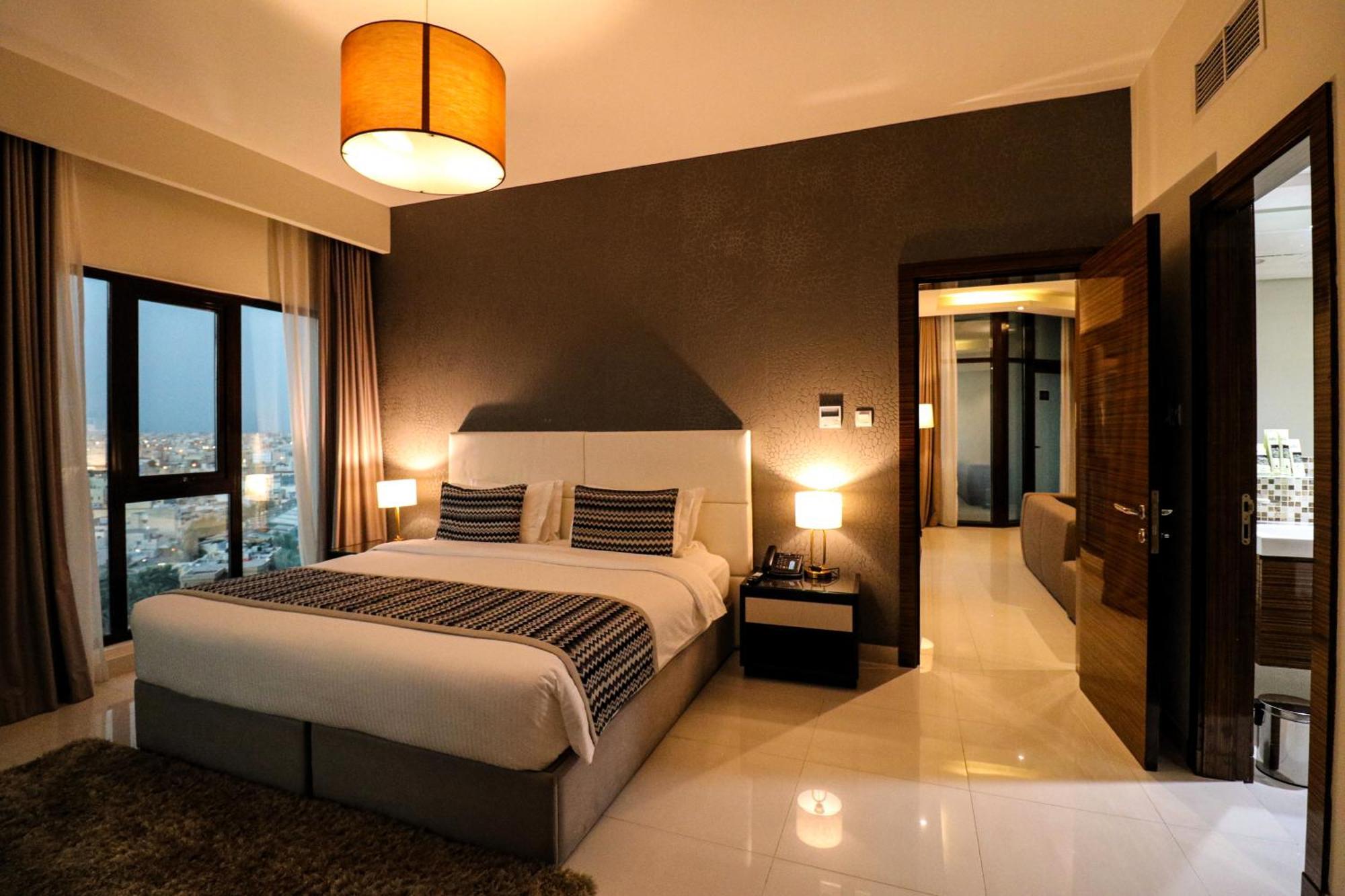 Lagoona Beach Luxury Resort And Spa Манама Экстерьер фото The photo depicts a modern hotel room or suite. It features a large bed with a white and gray color scheme, accented with decorative pillows. On either side of the bed, there are sleek bedside lamps providing warm lighting. The walls are a dark color