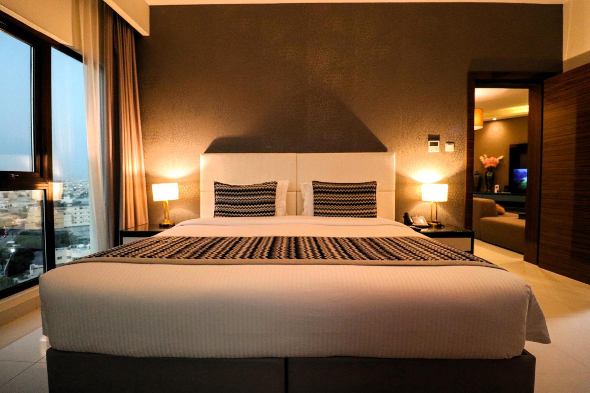 Lagoona Beach Luxury Resort And Spa Манама Экстерьер фото The photo depicts a modern hotel room featuring a large bed with a white comforter and two decorative pillows. The headboard is light-colored, and the walls are painted a dark hue. On either side of the bed, there are bedside tables with lamps. The r