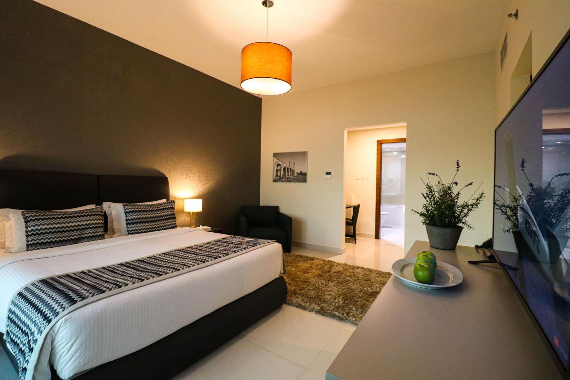 Lagoona Beach Luxury Resort And Spa Манама Экстерьер фото The photo shows a modern hotel room. It features a large bed with a neatly made white and dark-colored duvet. On either side of the bed, there are bedside tables with lamps. The wall behind the bed is painted in a dark color, creating a contrast with