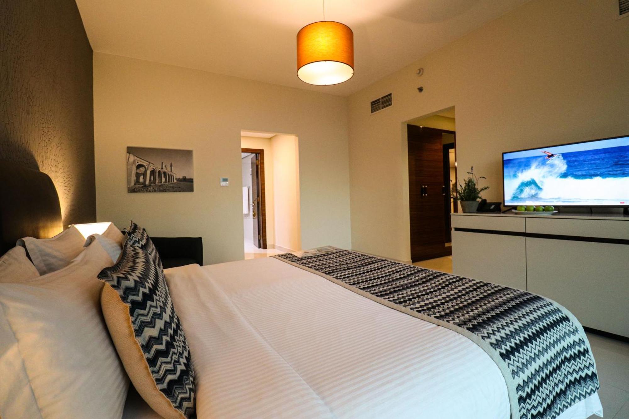 Lagoona Beach Luxury Resort And Spa Манама Экстерьер фото The photo shows a well-decorated hotel room. There is a large bed with white linens and decorative pillows featuring a zigzag pattern. On the wall beside the bed is a piece of artwork, and there is a modern television on a white furniture unit opposi