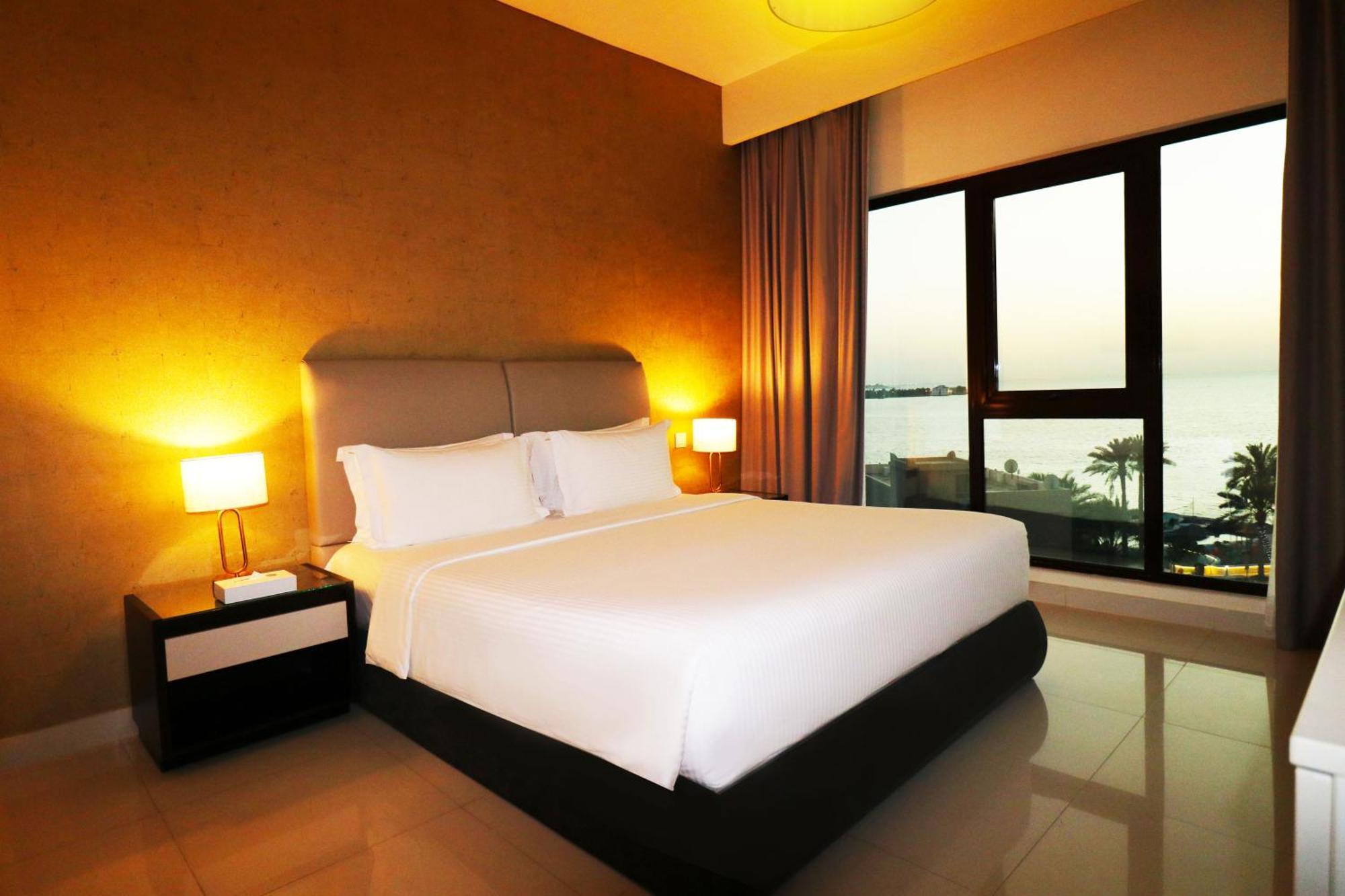 Lagoona Beach Luxury Resort And Spa Манама Экстерьер фото The photo shows a modern hotel bedroom with a large, comfortable bed dressed in white linens. The room features warm lighting, including bedside lamps, creating a cozy atmosphere. There are large windows with a view of the outdoors, possibly overlook