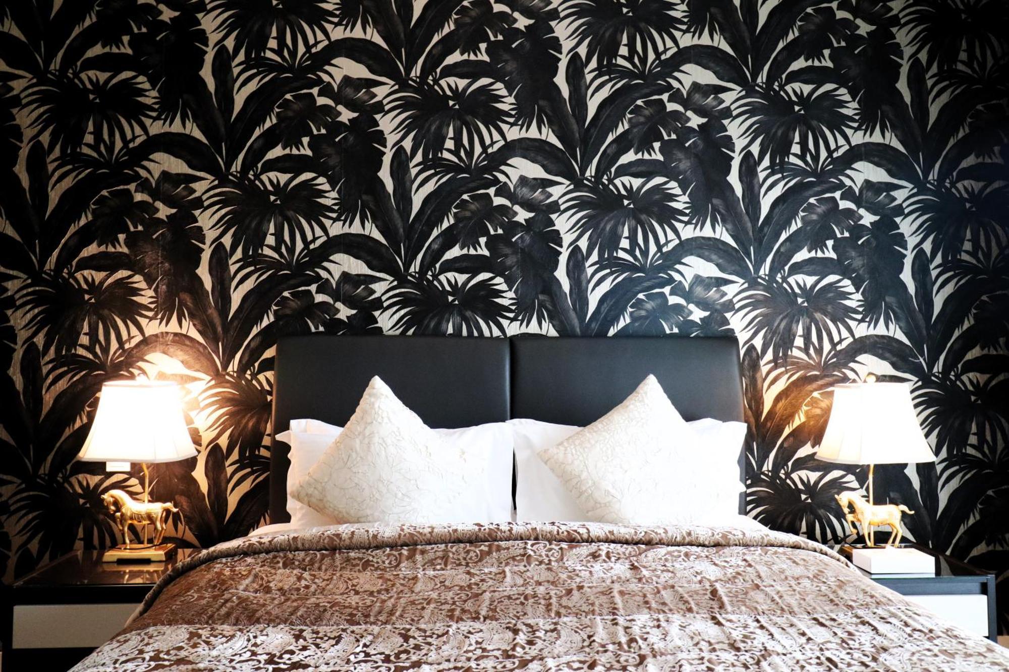 Lagoona Beach Luxury Resort And Spa Манама Экстерьер фото The photo depicts a stylish bedroom featuring a distinctive wall covered in a black and white tropical foliage wallpaper. The bed has a modern black headboard and is adorned with white pillows, creating a clean and elegant look. A textured brown thro