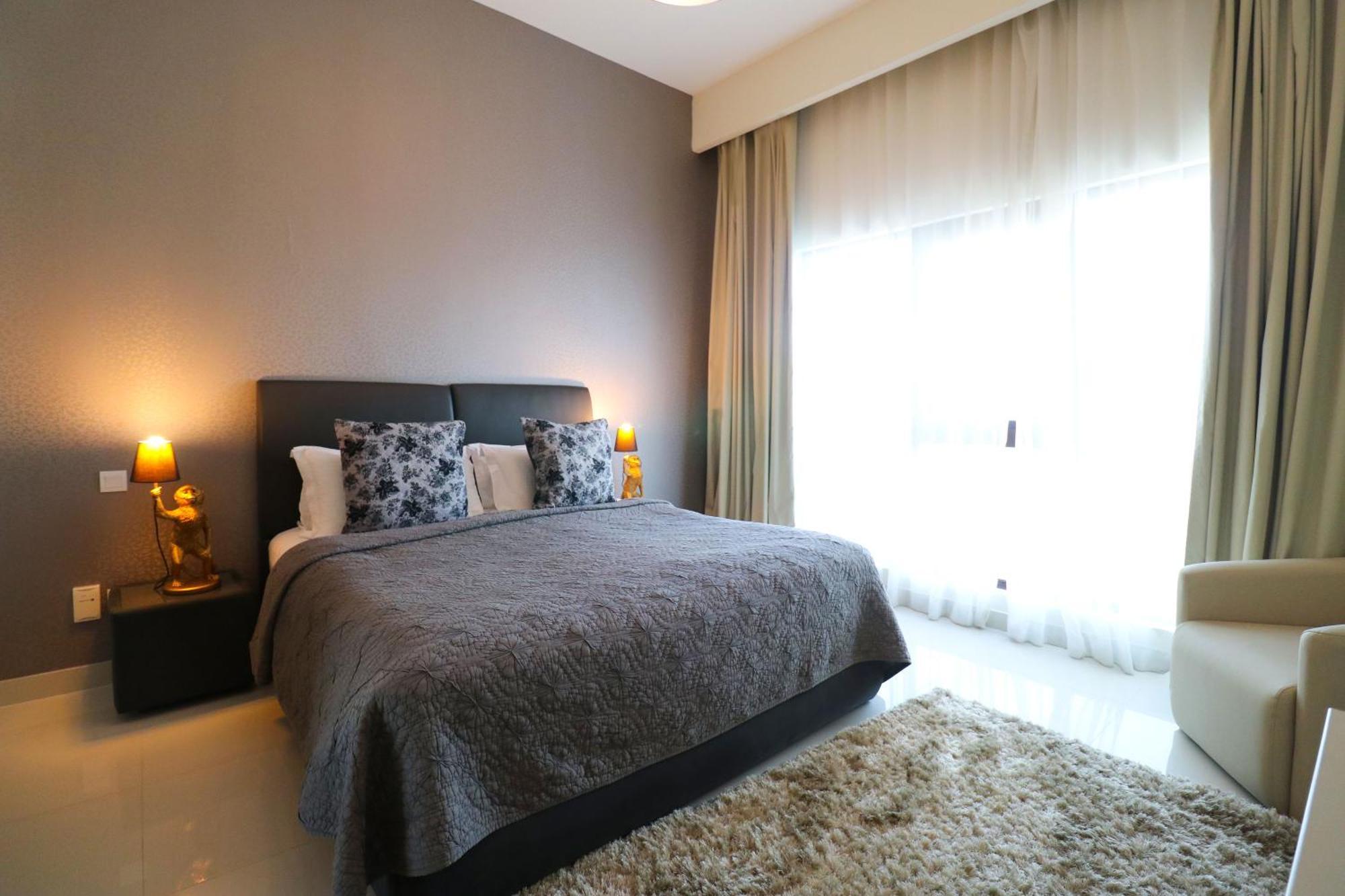 Lagoona Beach Luxury Resort And Spa Манама Экстерьер фото The photo shows a stylish bedroom with a modern design. It features a large bed with a dark gray bedspread and decorative pillows. On either side of the bed are two wooden nightstands, each with a lamp. There is a large window covered with light, flo
